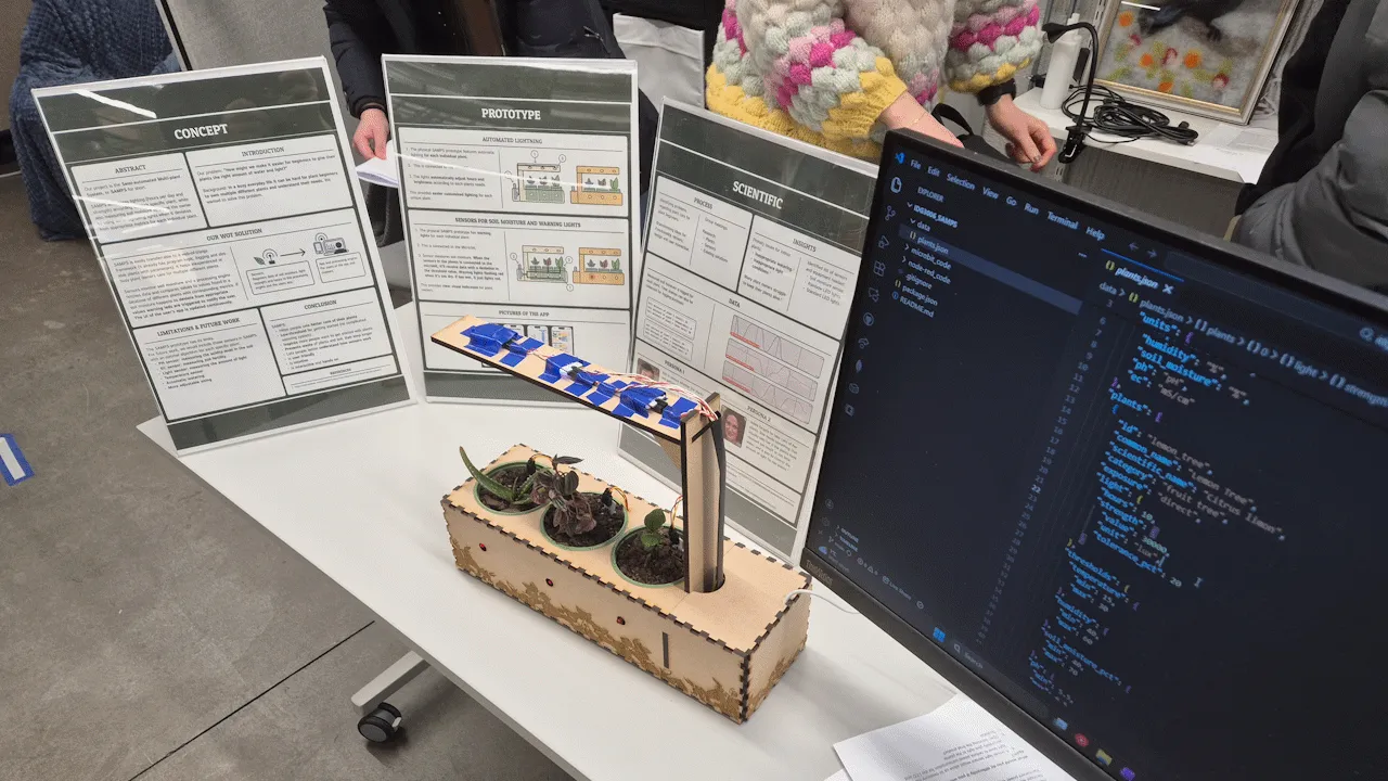Planteboks-prototypen med vanningsvarsler LED-er kan ses på bordet, sammen med informasjonsplakater og en skjerm med relatert kode.